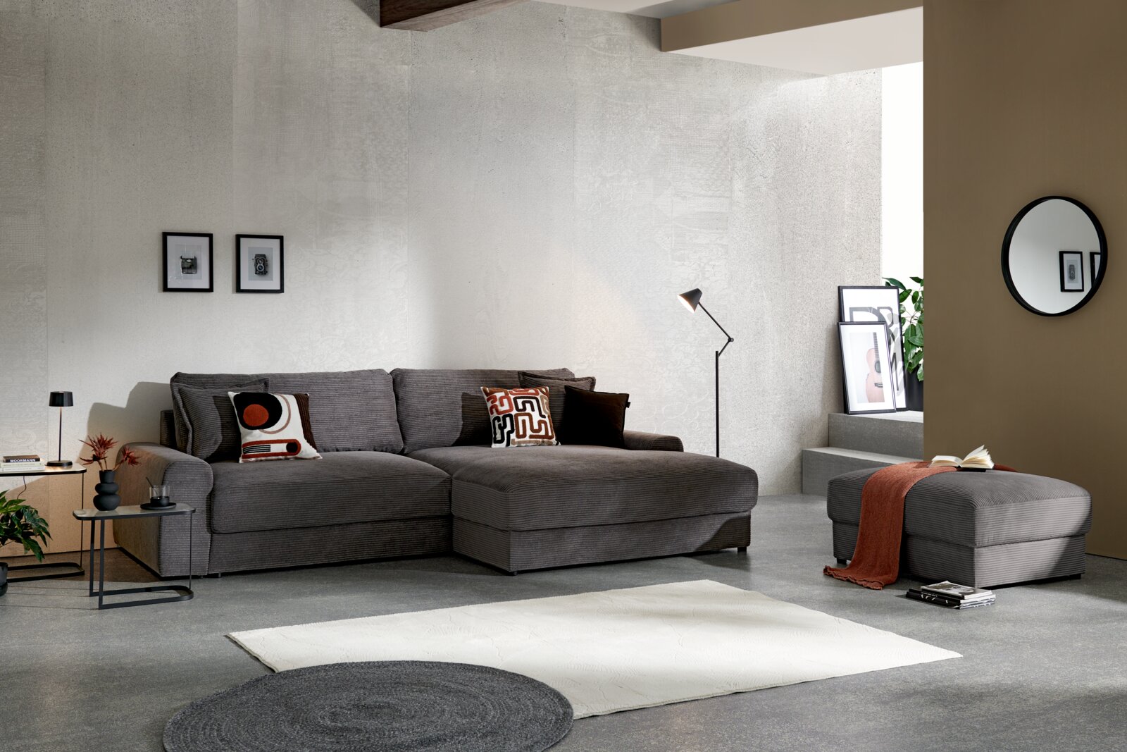 Modernes graues Cord-Ecksofa mit großer Ottomane und passenden Kissen im Wohnzimmer-Setting, Ansicht schräg von vorne.