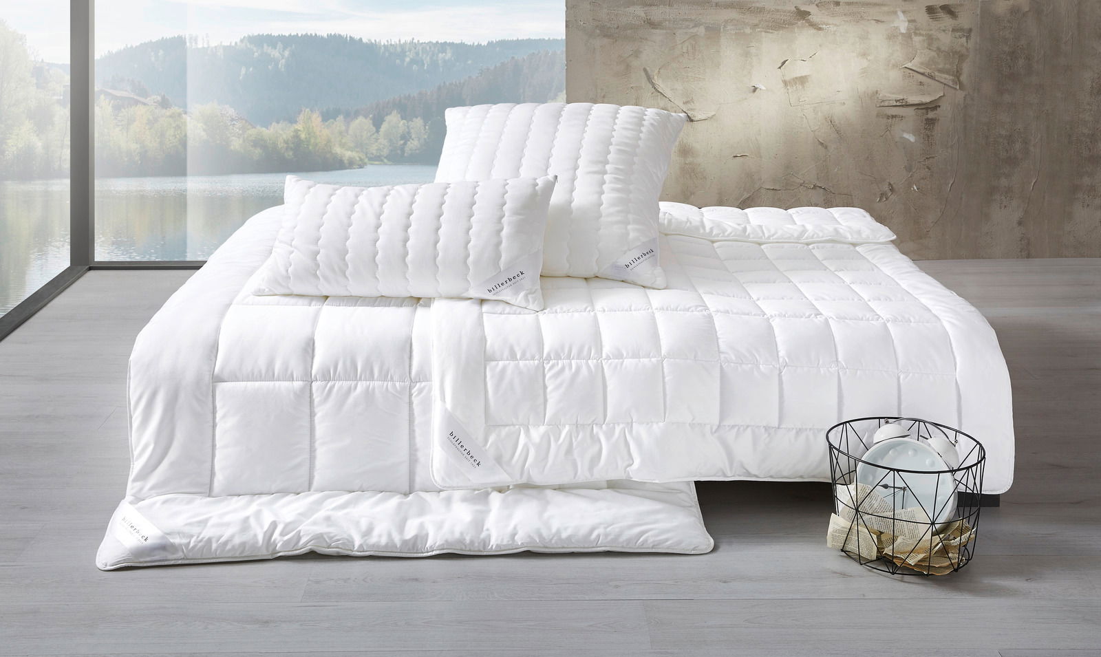billerbeck Faserdecke UNO ANTIGUA Blick auf ein Bett mit weißer Faserdecke und zwei weißen Kopfkissen in einem modernen Raum mit großem Fenster und Blick auf einen See.