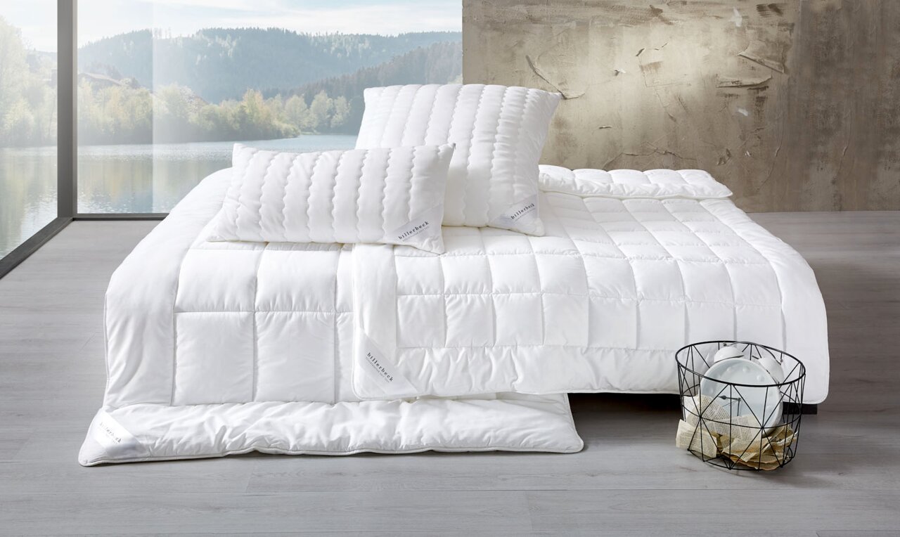 Blick auf ein Bett mit weißer Faserdecke und zwei weißen Kopfkissen in einem modernen Raum mit großem Fenster und Blick auf einen See.