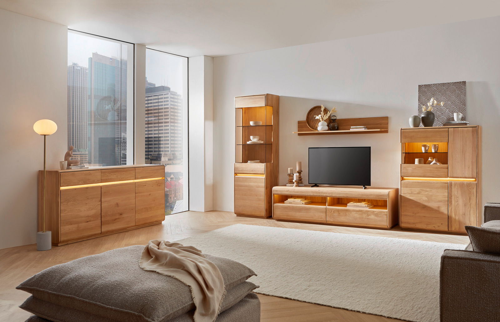 Wohnzimmer mit moderner Holzmöbel-Kombination, bestehend aus Sideboard, Vitrine, Wandboard und Highboard, mit LED-Beleuchtung, aus der Perspektive eines gemütlichen Wohnbereichs mit Blick auf die Stadt.