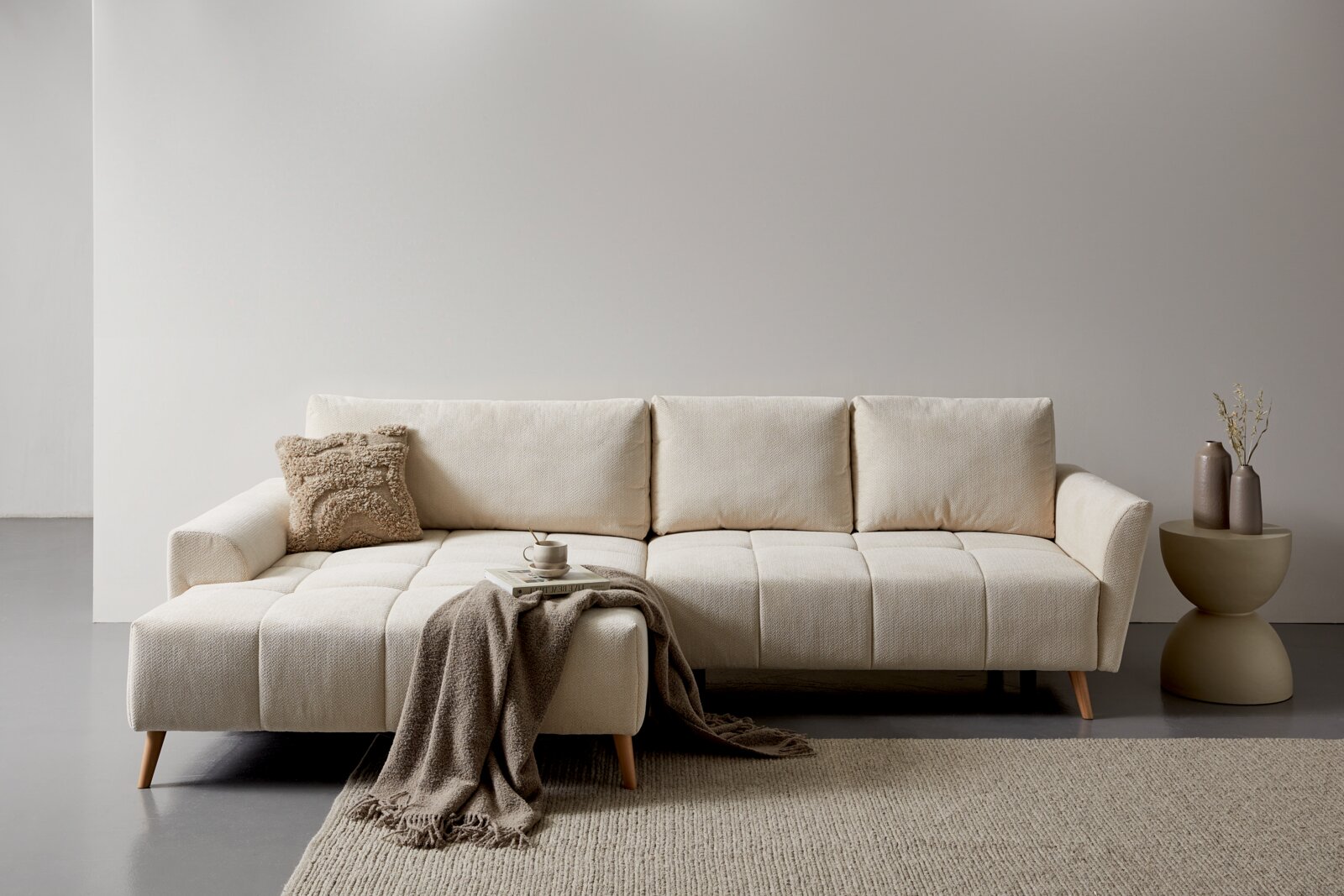Z2 Ecksofa ZELDA Modernes beiges Ecksofa mit Chaiselongue links, gesteppten Sitzflächen und Holzfüßen in minimalistischer Wohnzimmerszene, Frontansicht leicht schräg von vorn.