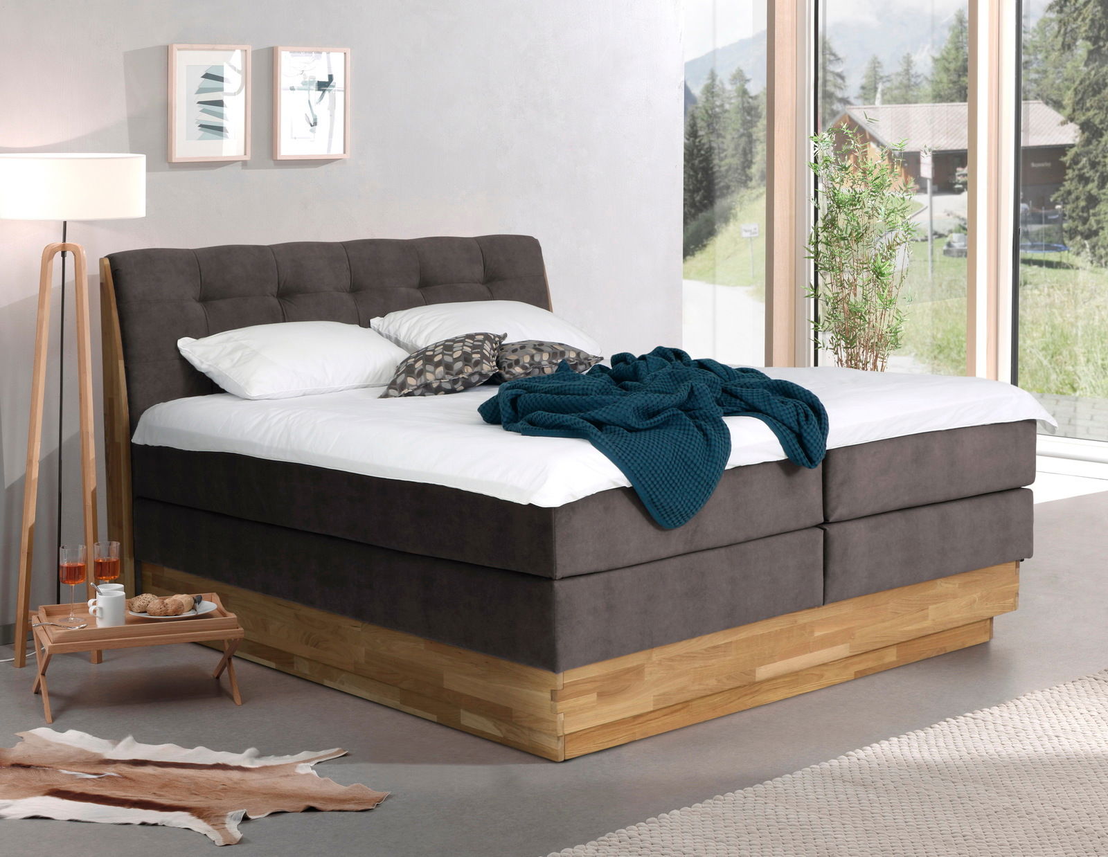 Modernes Boxspringbett in einem hellen Raum mit großen Fenstern, seitliche Perspektive.