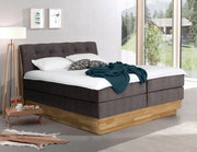 Modernes Boxspringbett in einem hellen Raum mit großen Fenstern, seitliche Perspektive.