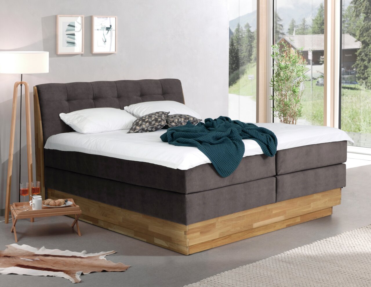 Modernes Boxspringbett in einem hellen Raum mit großen Fenstern, seitliche Perspektive.