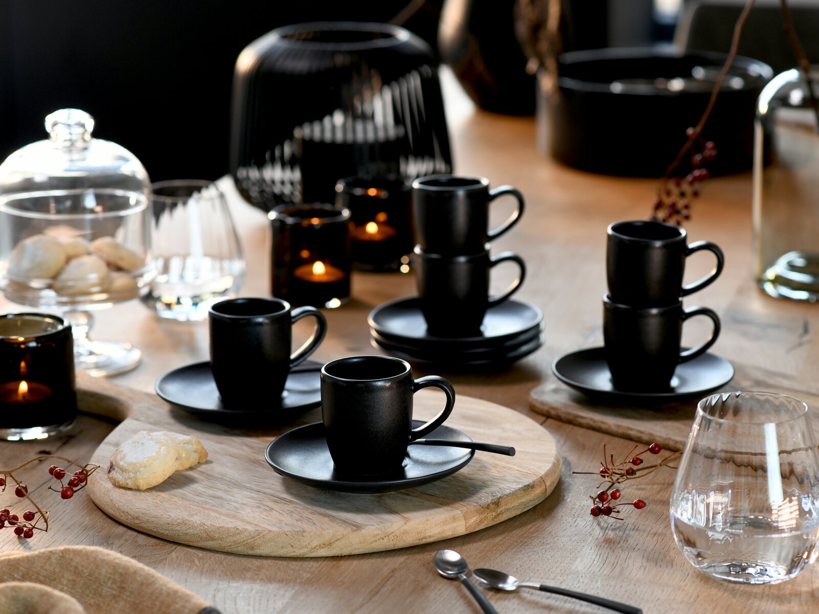 Schwarze Espressotassen mit Untertassen, teils gestapelt, auf einem gedeckten Holztisch mit Kerzen und Gläsern, fotografiert in leicht erhöhter, schräger Vorderansicht.