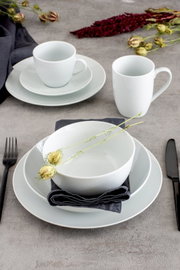 Schlichtes weißes Geschirr-Set mit tiefem Teller/Schüssel, Speisetellern und Tassen auf grauer Tischplatte, dekoriert mit dunkler Serviette und Trockenblumen, fotografiert in leicht schräger Draufsicht.