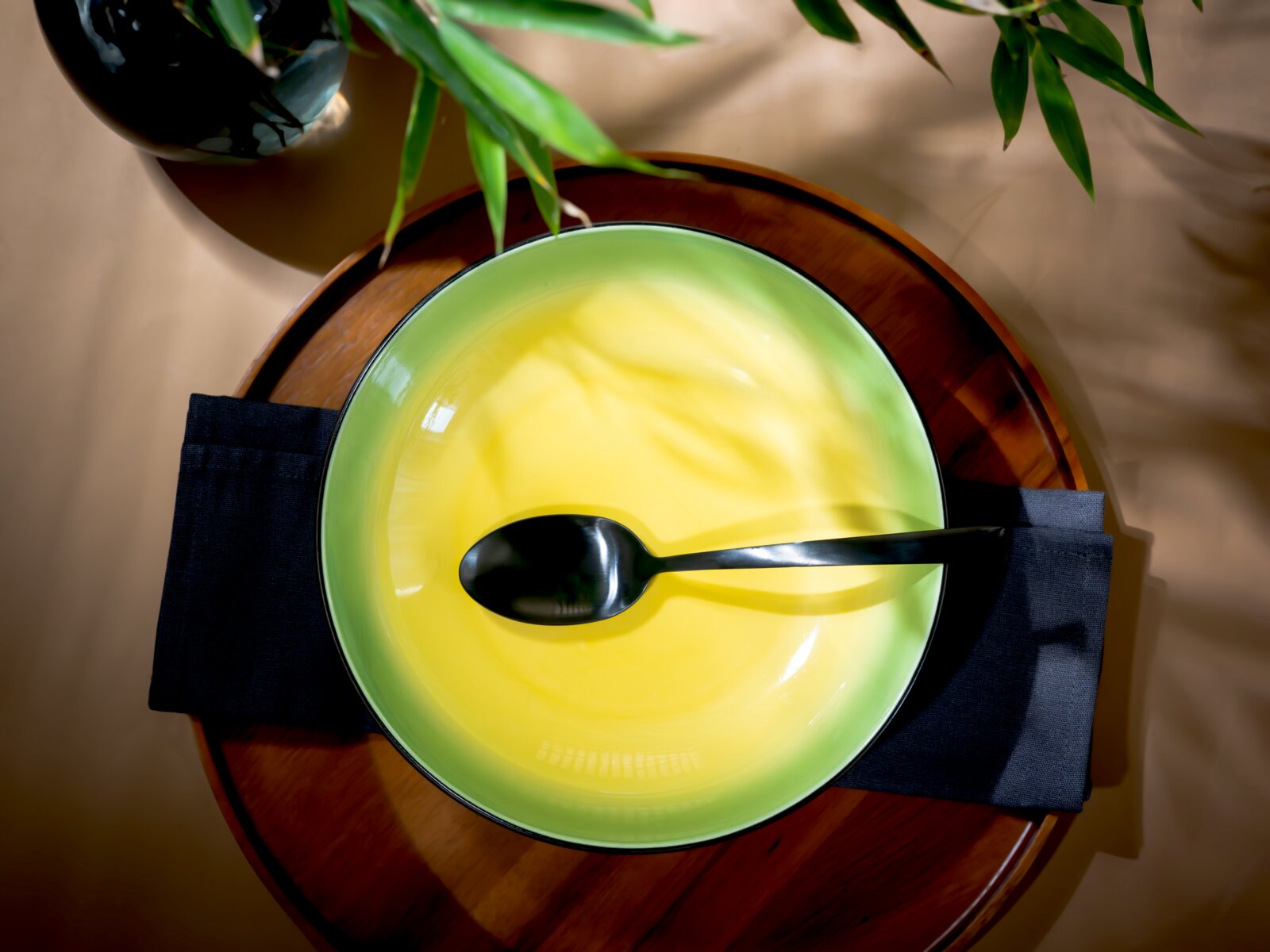 Grüne Keramikschale mit leuchtend gelber Cremesuppe und schwarzem Löffel auf einem runden Holztablett, aus der Vogelperspektive inszeniert mit grünen Pflanzenblättern.