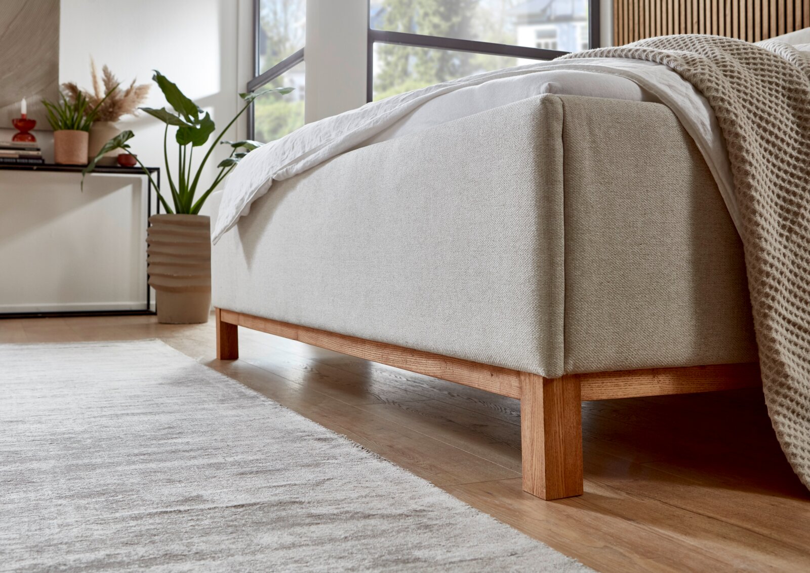 Zijaanzicht van een gestoffeerd bed met een lichte stoffen bekleding en houten poten in een moderne slaapkamer.