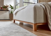 Zijaanzicht van een gestoffeerd bed met een lichte stoffen bekleding en houten poten in een moderne slaapkamer.