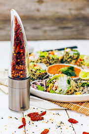 Chilischneider aus Edelstahl mit transparentem Behälter, gefüllt mit getrockneten Chilischoten, auf einem Tisch neben einem Teller mit asiatischem Essen. Perspektive: seitlich.