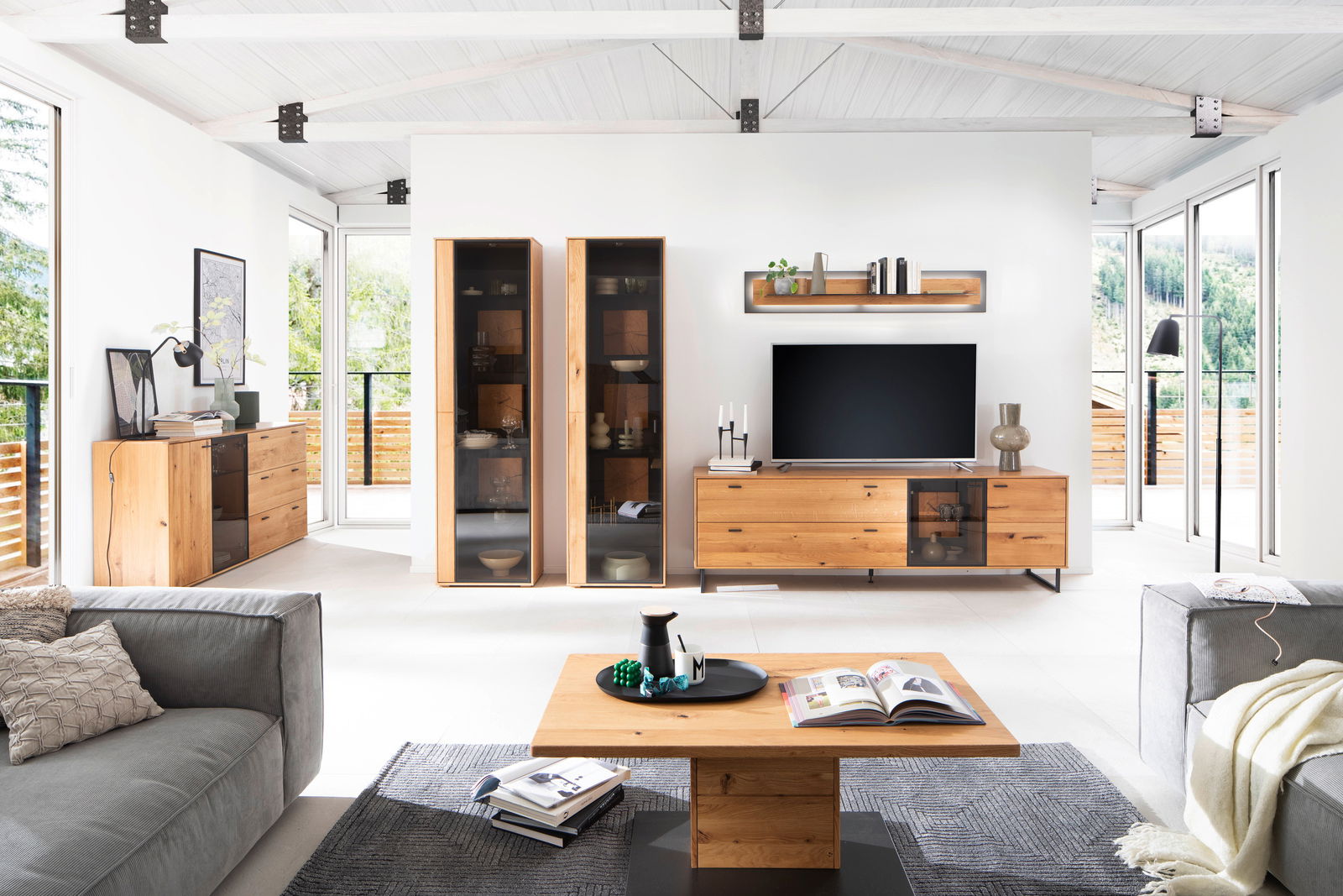 Modernes Wohnzimmer mit Wandboard in Holzoptik, zentral über einem Fernseher platziert, umgeben von stilvollen Möbeln und großen Fenstern mit Blick ins Grüne. Perspektive: Frontalansicht.