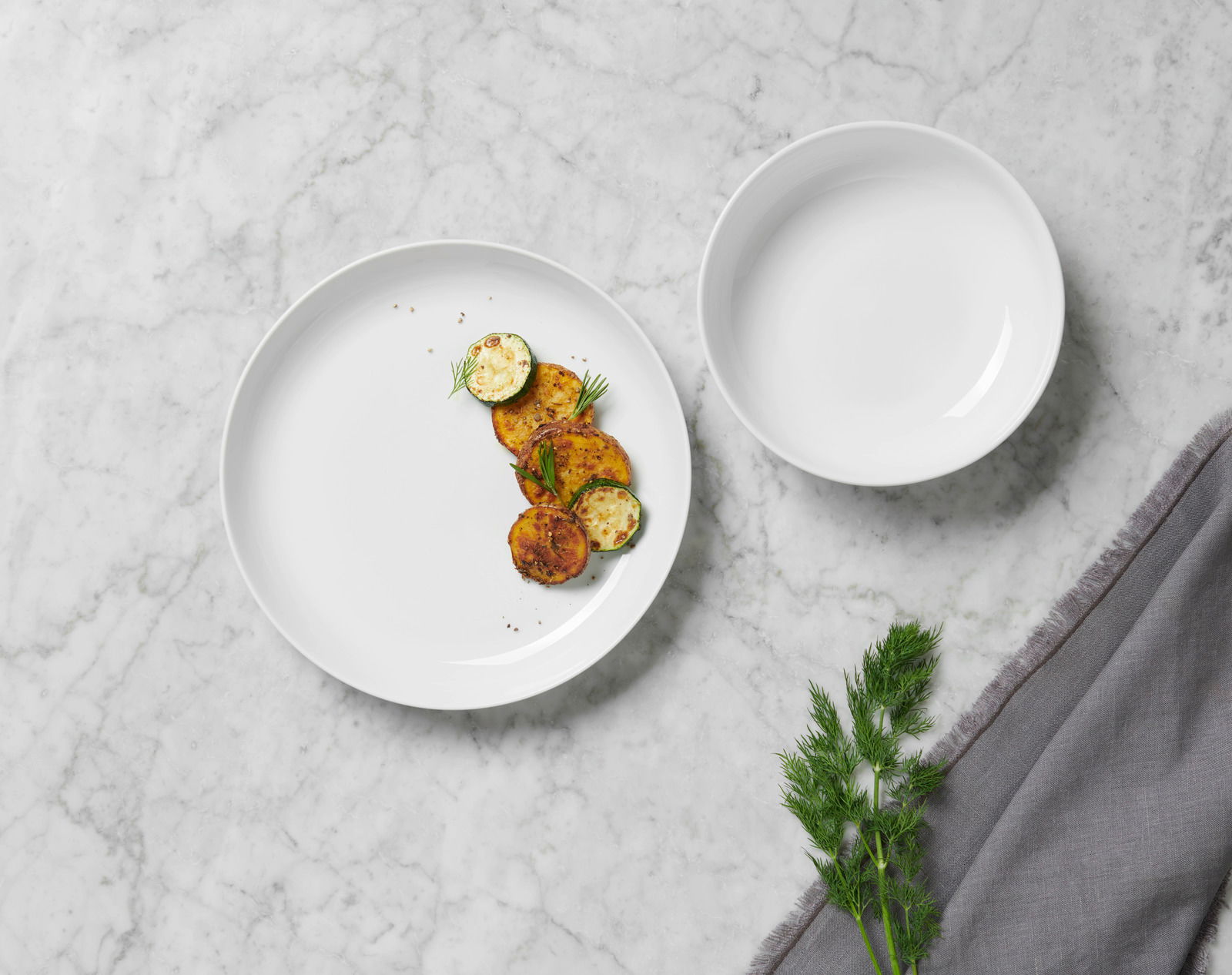 Vogelperspektive auf ein weißes Tafelservice mit einem Teller, der mit gebratenem Gemüse garniert ist, und einer leeren Schale auf einem Marmortisch.