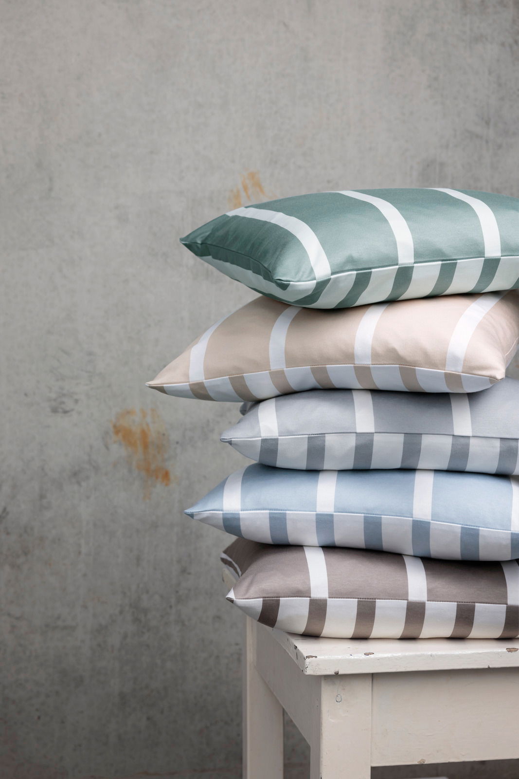 Ein Stapel von Kissenbezügen in verschiedenen Farben mit Streifenmuster, auf einem weißen Tisch vor einer grauen Wand von der Seite fotografiert.