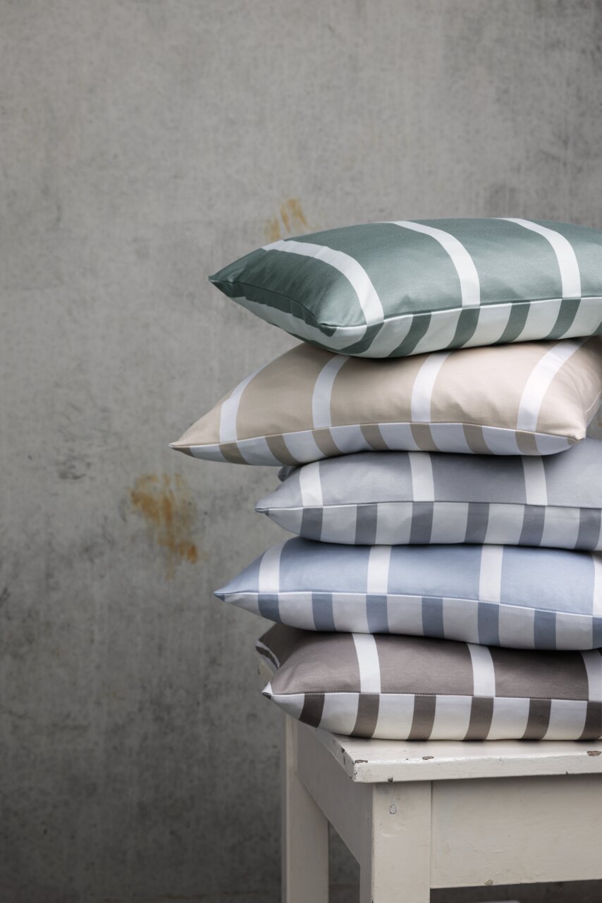 Ein Stapel von Kissenbezügen in verschiedenen Farben mit Streifenmuster, auf einem weißen Tisch vor einer grauen Wand von der Seite fotografiert.