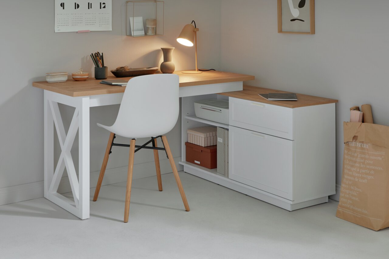 Moderner Eck-Schreibtisch in Holzoptik mit weißem Gestell, seitlicher Schubladenkommode und dekorativer Beleuchtung, aus der Perspektive eines gemütlichen Arbeitszimmers.