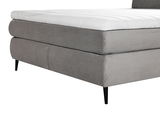 Nahaufnahme eines grauen Boxspringbetts mit gestepptem Stoffbezug und schwarzen Beinen, seitliche Perspektive