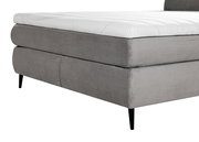 Nahaufnahme eines grauen Boxspringbetts mit gestepptem Stoffbezug und schwarzen Beinen, seitliche Perspektive