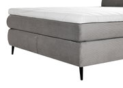 Nahaufnahme eines grauen Boxspringbetts mit gestepptem Stoffbezug und schwarzen Beinen, seitliche Perspektive