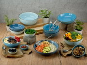 Keramik-Schüsselset in Blau- und Türkistönen mit gesprenkelter Glasur, dekorativ auf Holztisch arrangiert und mit Bowls, Obst, Nüssen sowie Besteck gestylt; Perspektive: leicht von oben in schräger Frontansicht.