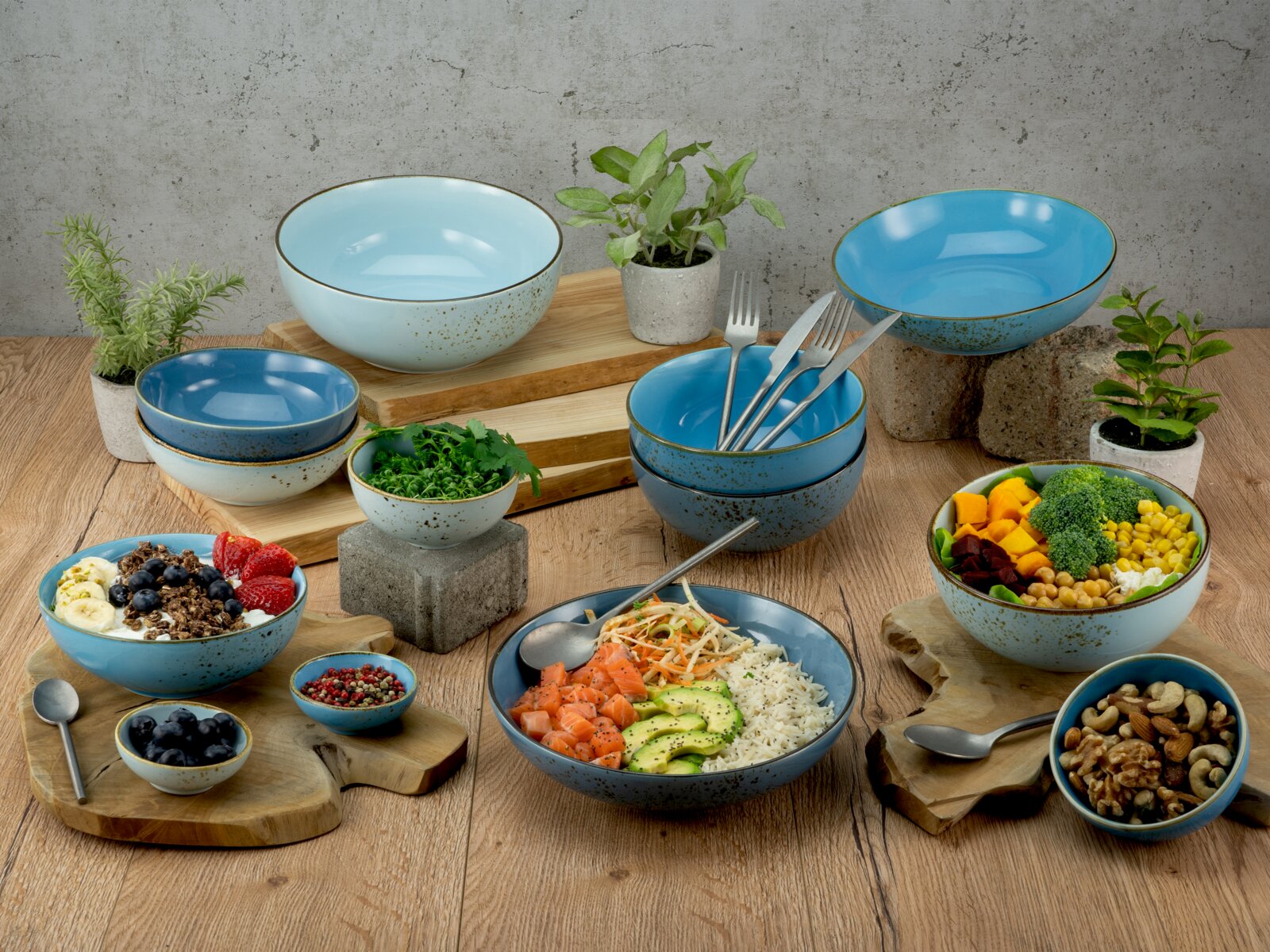 Keramik-Schüsselset in Blau- und Türkistönen mit gesprenkelter Glasur, dekorativ auf Holztisch arrangiert und mit Bowls, Obst, Nüssen sowie Besteck gestylt Perspektive: leicht von oben in schräger Frontansicht.