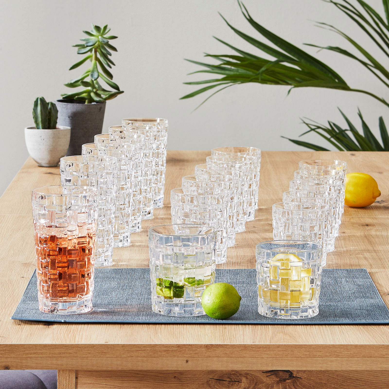 Becherset Bossa Nova 18-tlg. auf einem Holztisch, mit Getränken und Limetten, aus seitlicher Perspektive fotografiert.