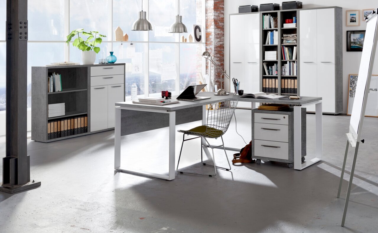 Moderner Eck-Schreibtisch in einem hellen Büro mit großen Fenstern, ausgestattet mit einem Rollcontainer und einem Drahtstuhl, aus der Perspektive von vorne rechts betrachtet.