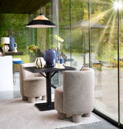 Zwei moderne, beige Sessel mit runden Formen stehen an einem schwarzen Tisch in einem lichtdurchfluteten Raum mit großen Glasfenstern. Die Perspektive zeigt die Sessel von der Seite.