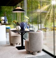 Zwei moderne, beige Sessel mit runden Formen stehen an einem schwarzen Tisch in einem lichtdurchfluteten Raum mit großen Glasfenstern. Die Perspektive zeigt die Sessel von der Seite.