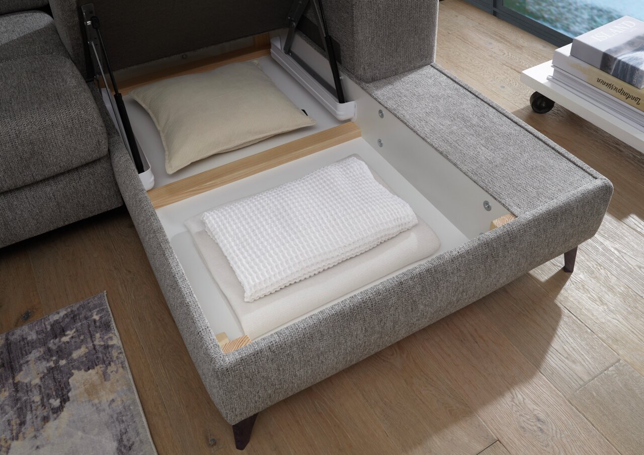 Perspektive von oben auf eine geöffnete graue Rundecke mit Stauraum, in dem ein Kissen und eine gefaltete Decke liegen.