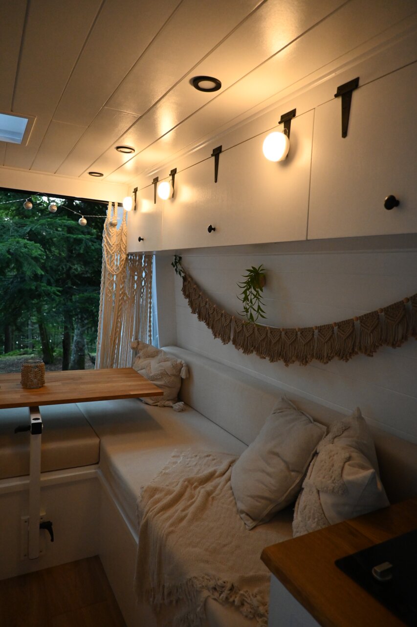 Gezellige inrichting van een busje met een lichte zitbank, een houten tafel en sfeervolle wandlampen, gefotografeerd vanuit het interieur, schuin naar voren in de richting van het raam.