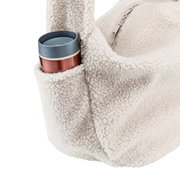 Close-up van een crèmekleurige tas van teddyfleece met ritssluiting en een buitenvak waarin een beker zit, gefotografeerd vanuit een zijdelingse hoek tegen een witte achtergrond.