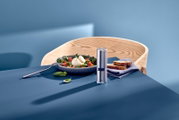 Edelstahl-Pfeffermühle mit transparentem Mittelteil auf blauem Tisch neben Salatschale und Brot, aufgenommen in leichter Seitenansicht von vorne