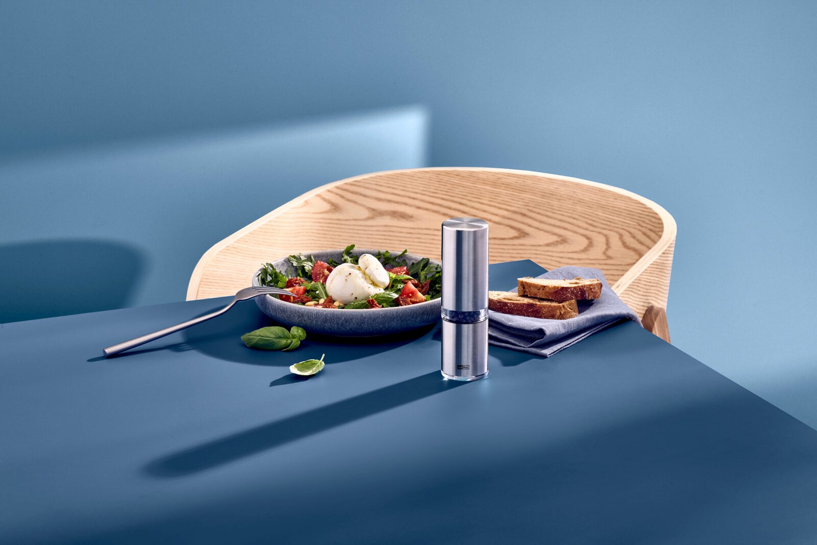 Edelstahl-Pfeffermühle mit transparentem Mittelteil auf blauem Tisch neben Salatschale und Brot, aufgenommen in leichter Seitenansicht von vorne