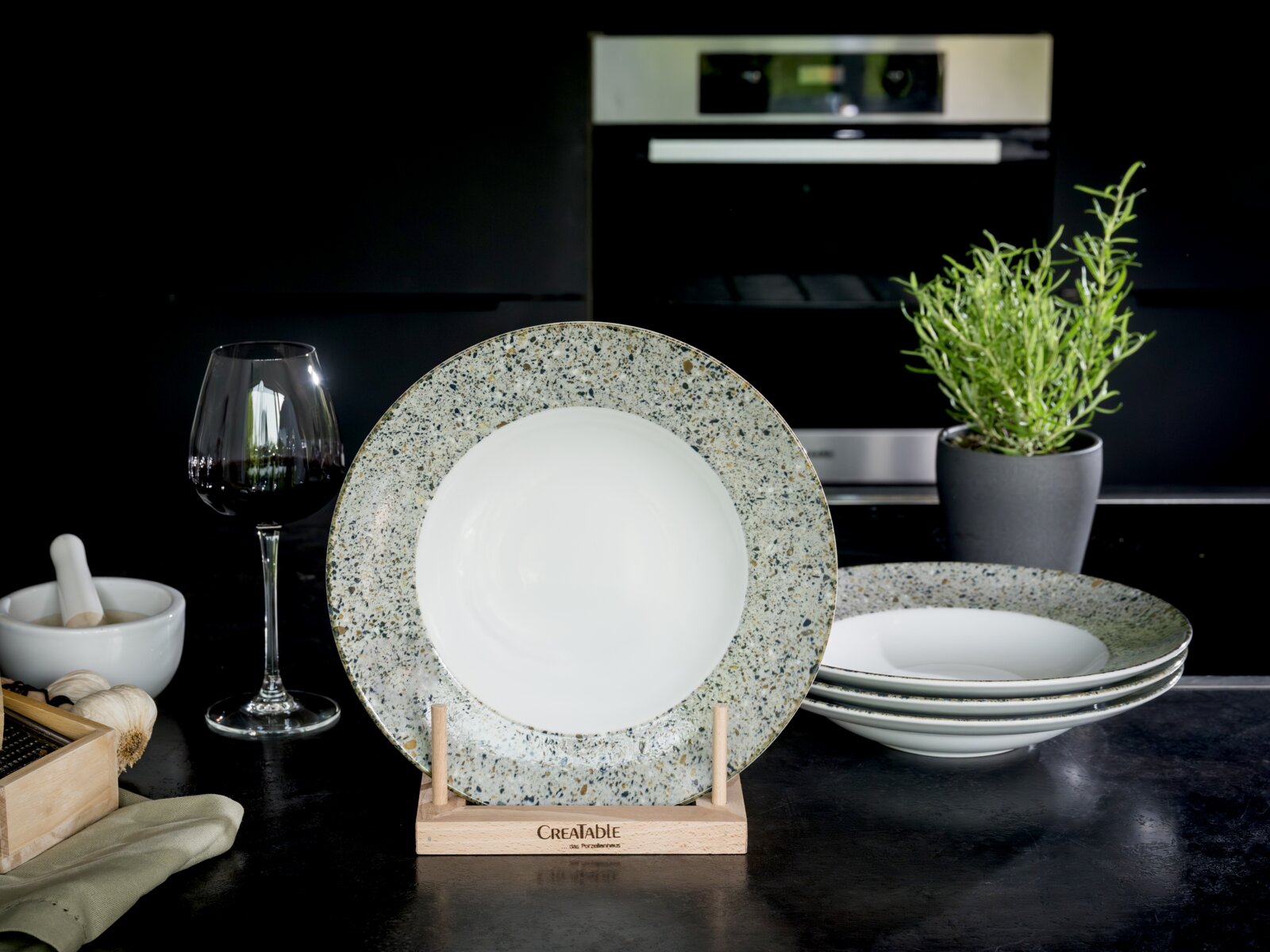 Creatable Pastateller 4-tlg STONE COLLECTION Speiseteller aus Porzellan mit weißer Mitte und gesprenkeltem, steinoptikartigem Rand auf Holzständer, frontale Ansicht auf Küchenarbeitsplatte; im Hintergrund weitere Teller, Rotweinglas und Kräutertopf.