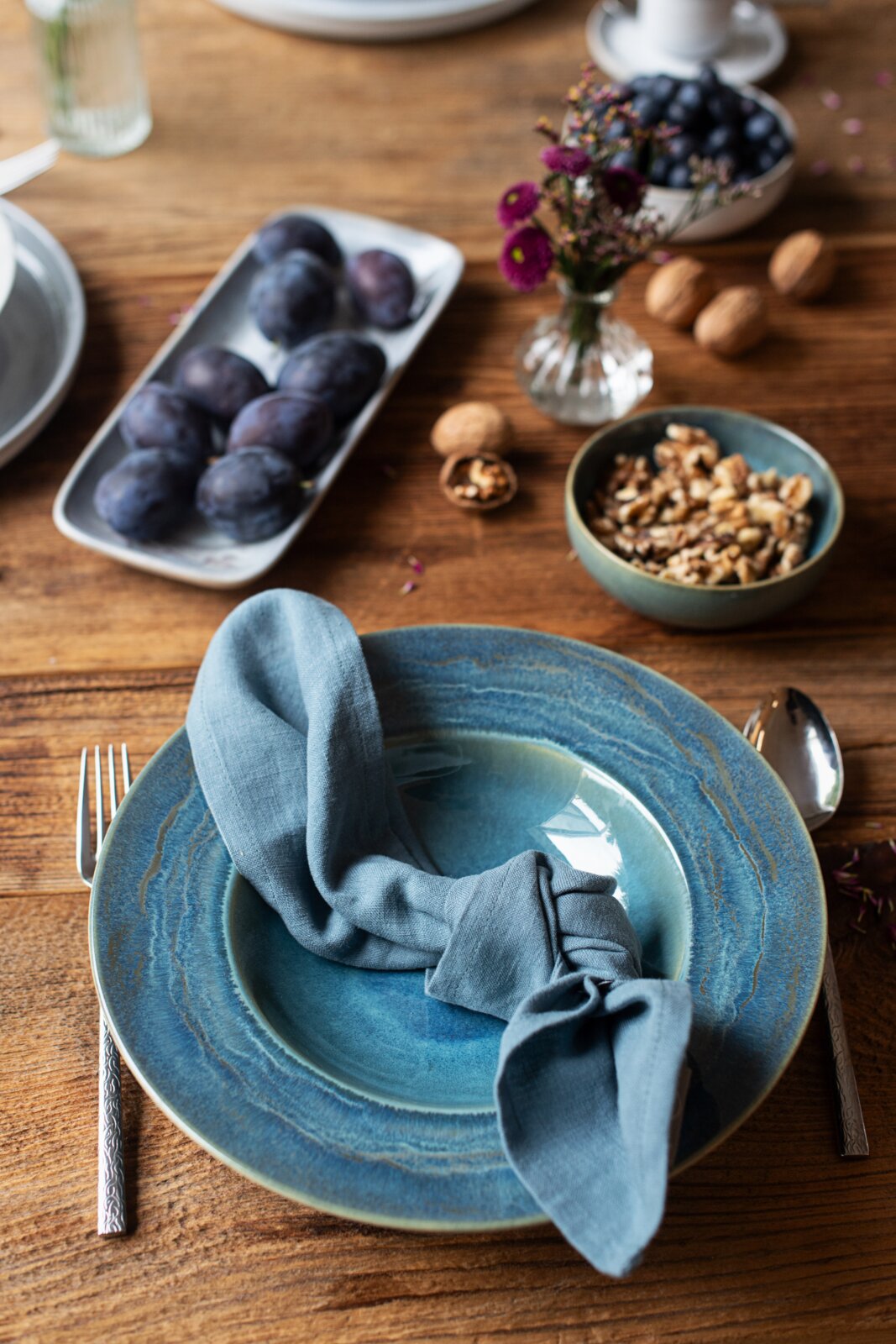 Schräg von oben fotografiertes Tischgedeck mit blau glasiertem Keramikteller und hellblauer Stoffserviette, umgeben von Besteck, Schale mit Walnüssen, Pflaumen und Trauben auf rustikalem Holztisch.
