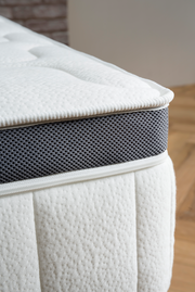 Nahaufnahme einer Matratze mit weißem, strukturiertem Stoff und grauem, atmungsaktivem Seitenstreifen. Perspektive von der Ecke der Matratze.