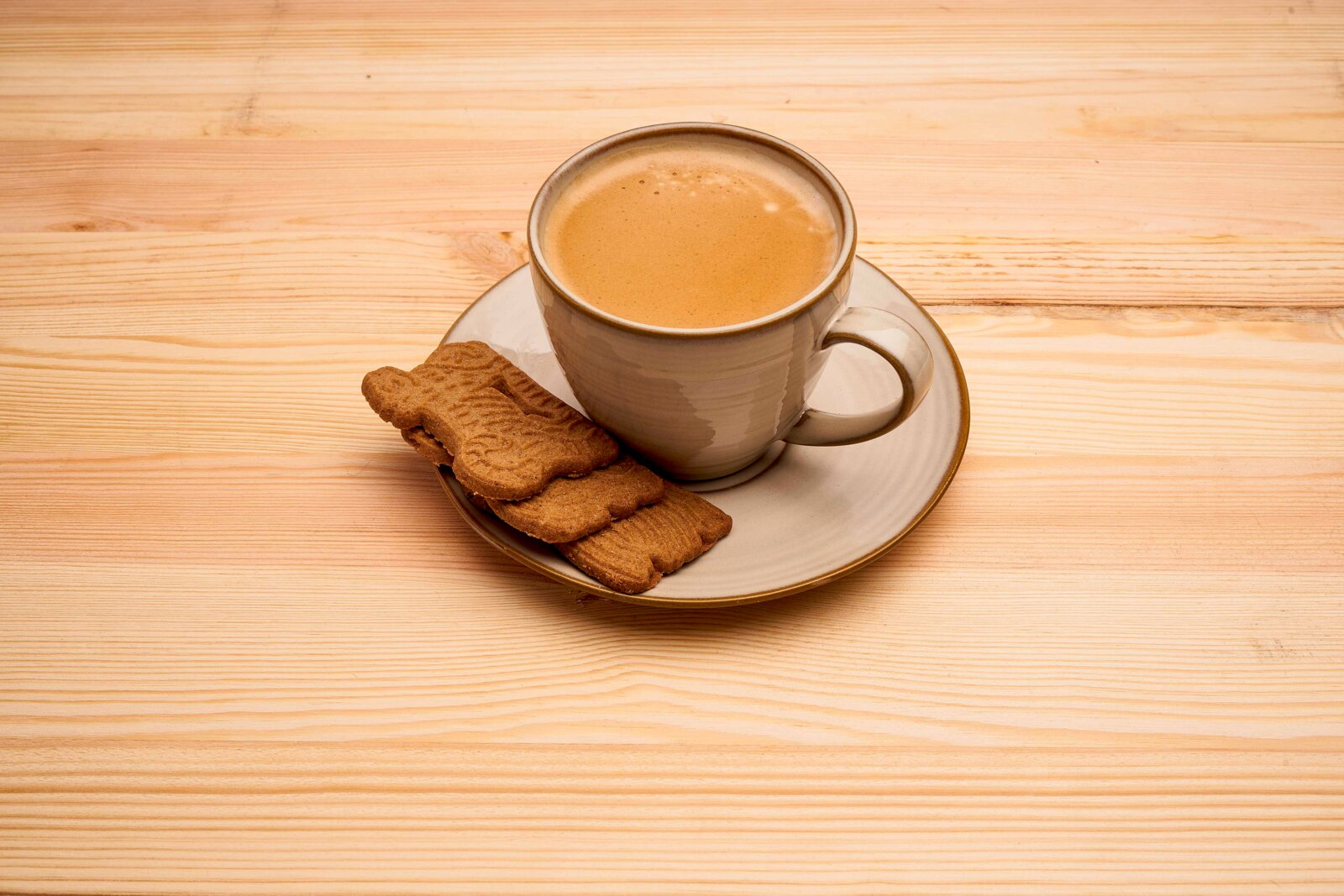Graue Keramiktasse mit Kaffee auf Untertasse und zwei Keksen auf hellem Holztisch, leicht von oben in schräger Draufsicht fotografiert