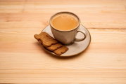 Graue Keramiktasse mit Kaffee auf Untertasse und zwei Keksen auf hellem Holztisch, leicht von oben in schräger Draufsicht fotografiert