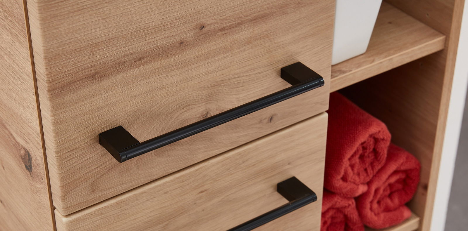 Nahaufnahme eines Midischranks aus Holz mit schwarzen Griffen, seitliche Perspektive, zeigt geöffnete Regale mit roten Handtüchern.
