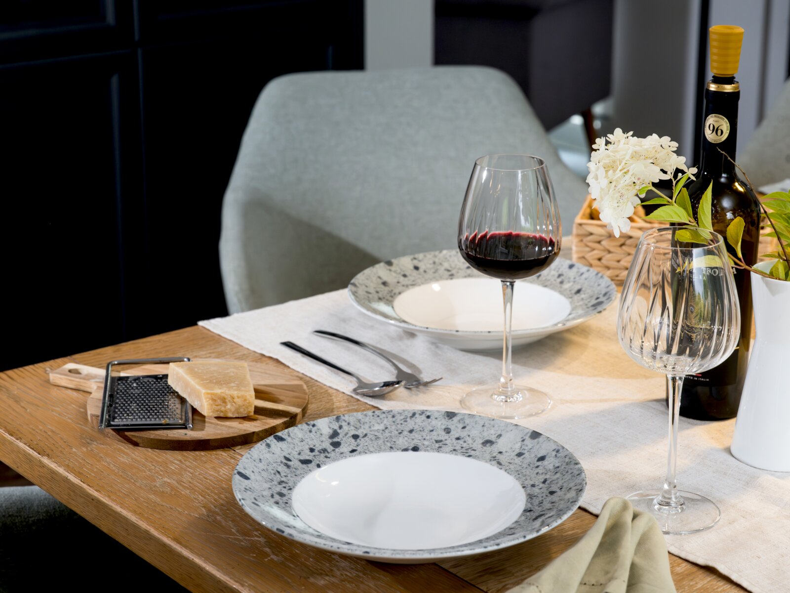 Gedeckter Esstisch mit gesprenkelten Keramiktellern, zwei Weingläsern (eins mit Rotwein), Weinflasche, Käse und Reibe auf Holzbrett sowie Blumenarrangement, aufgenommen aus schräger Seitenansicht auf Tischhöhe