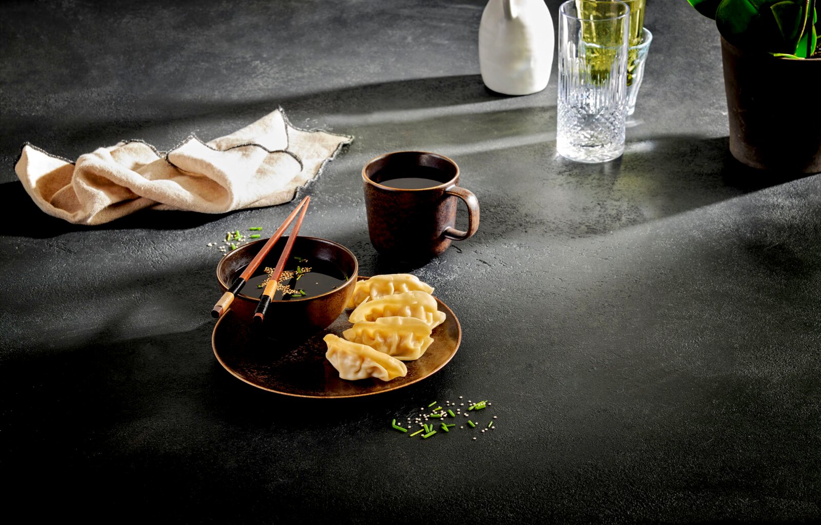 Rustikales dunkelbraunes Keramik-Geschirrset mit Schale, Teller und Tasse, dazu Essstäbchen und Dumplings auf dunkler Steinoptik-Tischplatte, schräg von oben fotografiert.