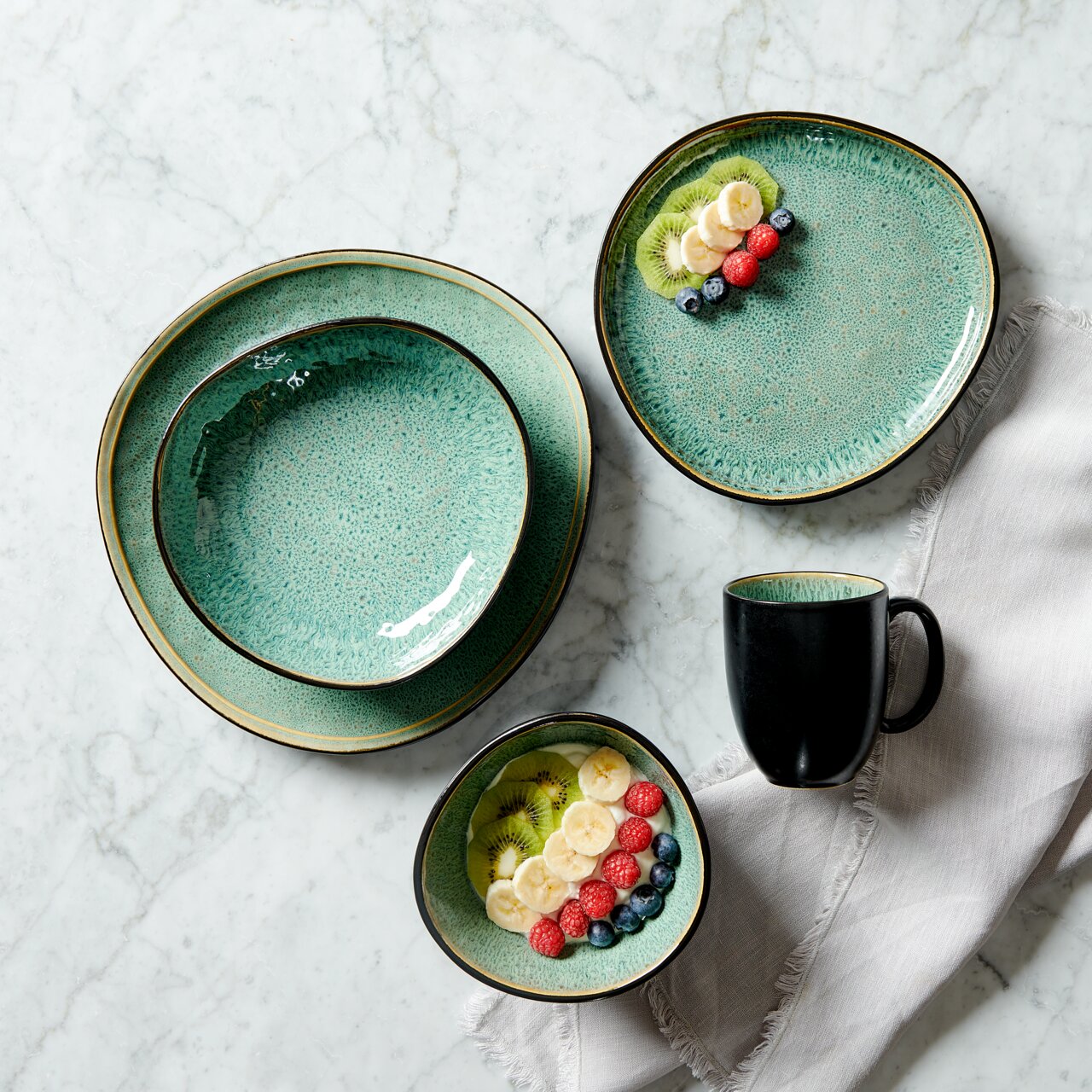 Kombiservice 10-teilig in grüner Keramikoptik, bestehend aus Tellern, Schalen und einer Tasse, von oben fotografiert auf einem Marmortisch mit Früchten dekoriert.