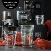 Twee kelkglaasjes in close-up, omringd door verpakkingsmateriaal, met een koffiezetapparaat op de achtergrond. Perfect als cadeau-idee.