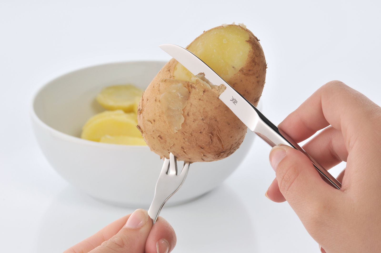 Nahaufnahme eines Pellkartoffel-Sets mit Messer und Gabel, die eine Kartoffel schälen, im Hintergrund eine Schale mit Kartoffelscheiben.