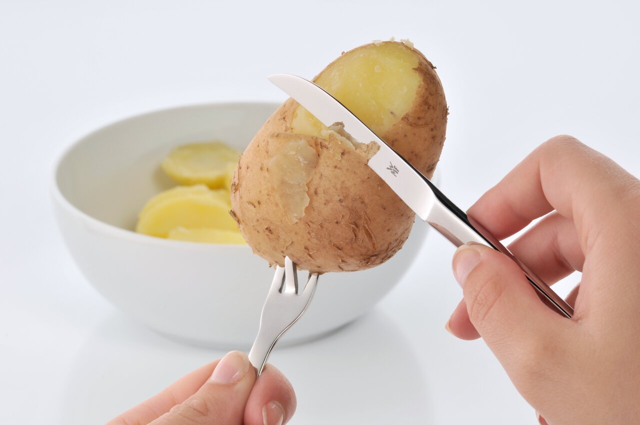 Nahaufnahme eines Pellkartoffel-Sets mit Messer und Gabel, die eine Kartoffel schälen, im Hintergrund eine Schale mit Kartoffelscheiben.