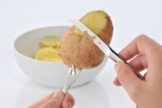 Nahaufnahme eines Pellkartoffel-Sets mit Messer und Gabel, die eine Kartoffel schälen, im Hintergrund eine Schale mit Kartoffelscheiben.