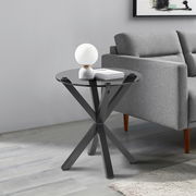 Moderner Beistelltisch mit schwarzer Glasplatte und gekreuztem Metallgestell, seitliche Perspektive neben einem grauen Sofa.