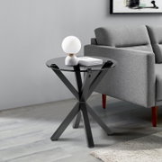 Moderner Beistelltisch mit schwarzer Glasplatte und gekreuztem Metallgestell, seitliche Perspektive neben einem grauen Sofa.