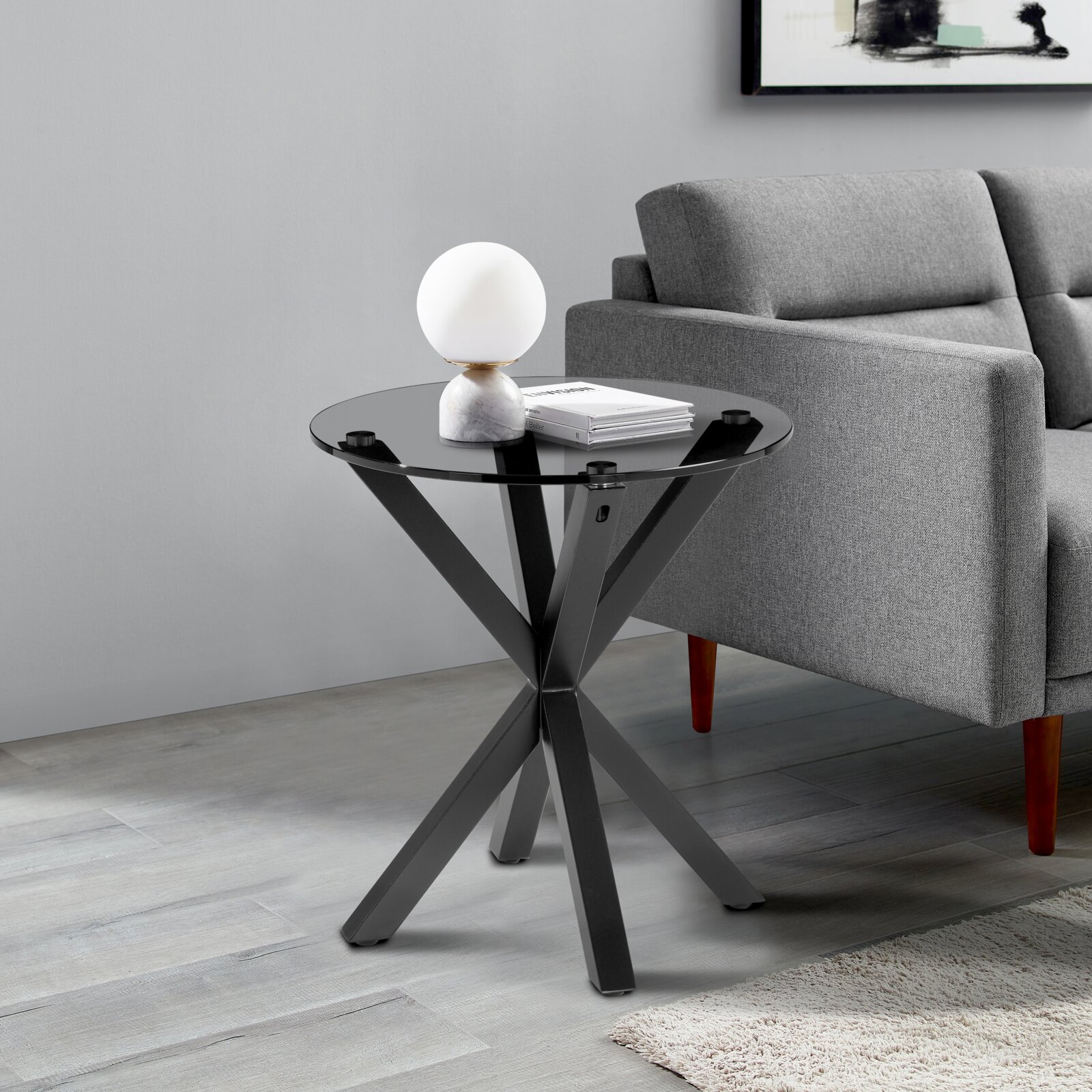 Moderner Beistelltisch mit schwarzer Glasplatte und gekreuztem Metallgestell, seitliche Perspektive neben einem grauen Sofa.