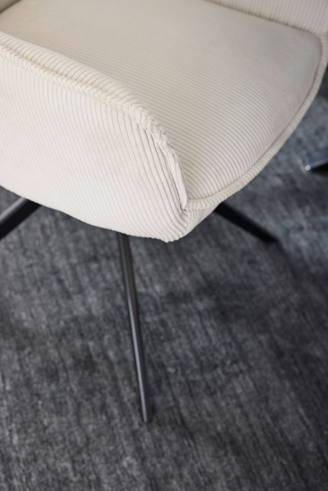 Nahaufnahme eines cremefarbenen Armlehnstuhls mit geripptem Stoffbezug und schwarzen Metallbeinen, von oben betrachtet.